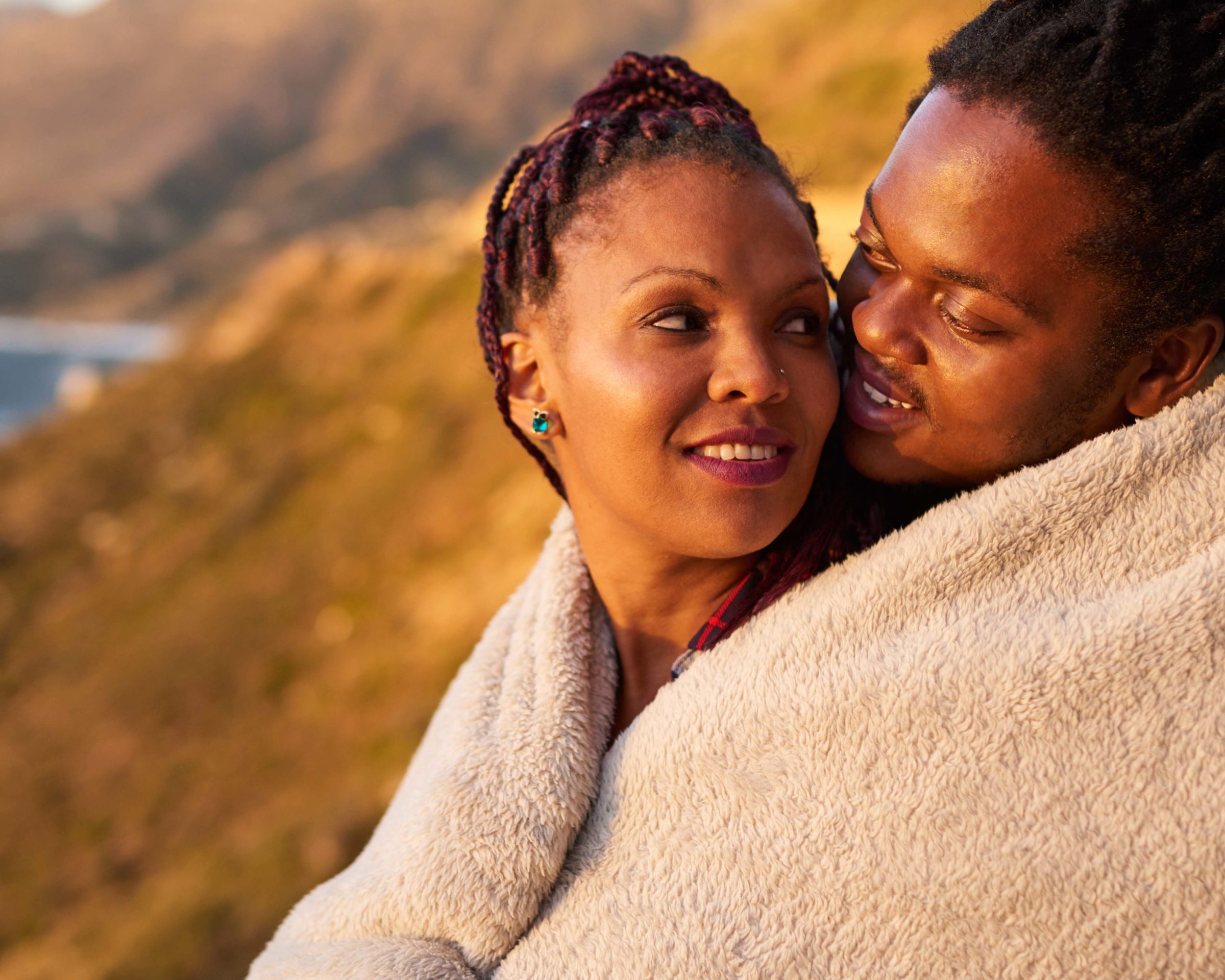Couple keeping warm under a blanket outdoors, with a stunning view of mountains and the ocean behind them, they enjoy an inimate moment with each other by showing their affection for one and other.